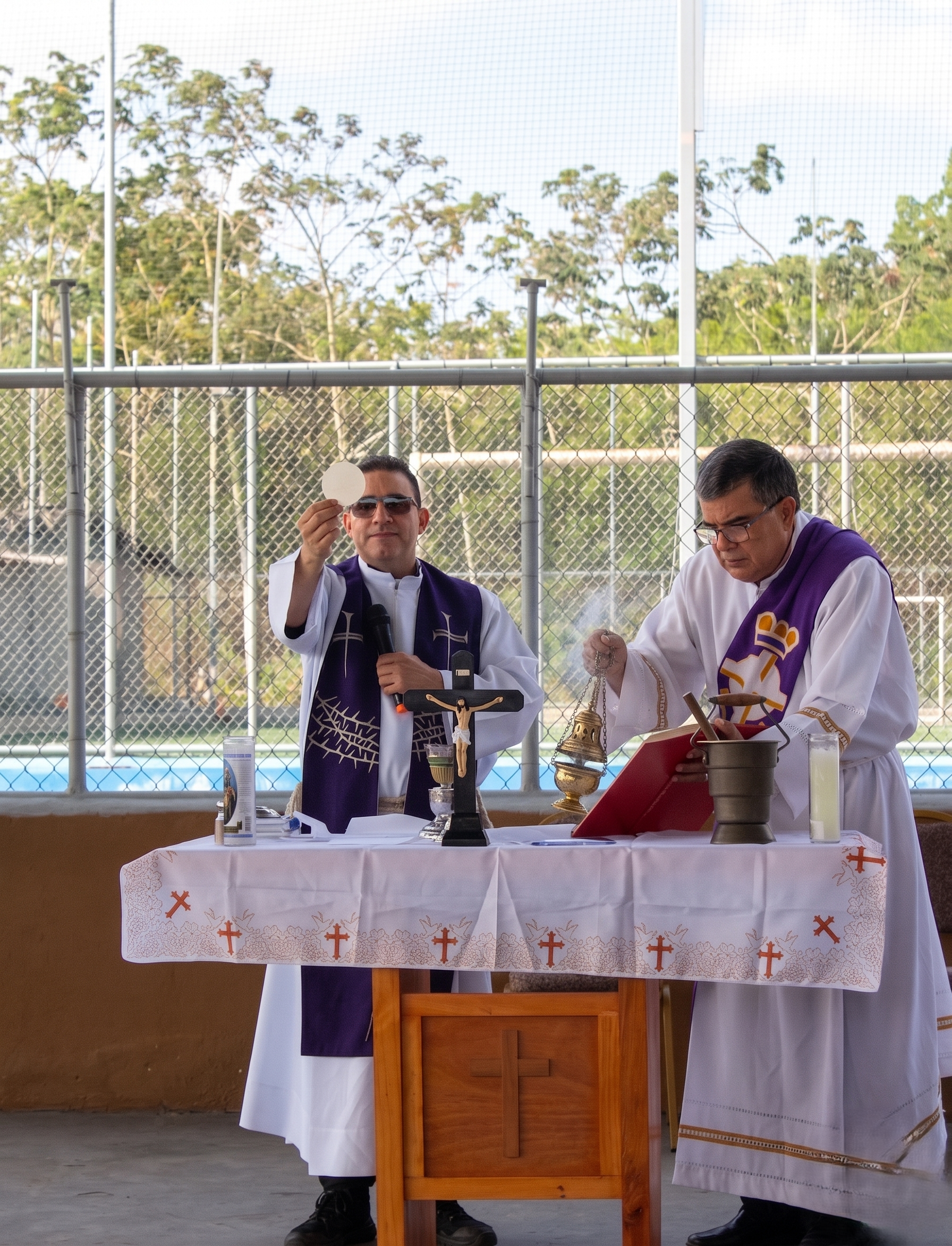 Imagen de dos sacerdotes en una ceremonia religiosa al aire libre, con mesa, cruces y objetos religiosos, frente a una cerca de malla y árboles.