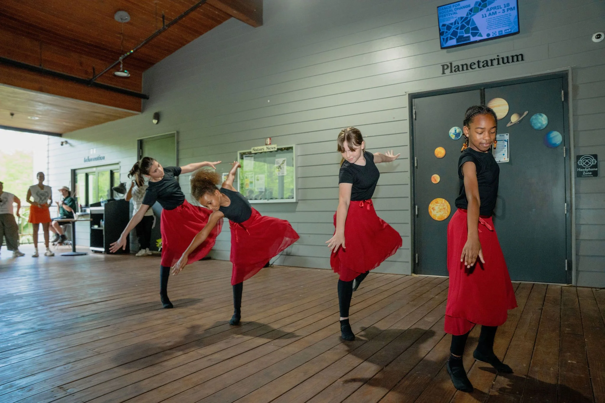 A group of young girls performing a dance routine in a space with a sign that reads 'Planetarium'. The girls are dressed in black tops and red skirts, and are doing a synchronized dance with raised arms and bend-forward postures, on a wooden floor.