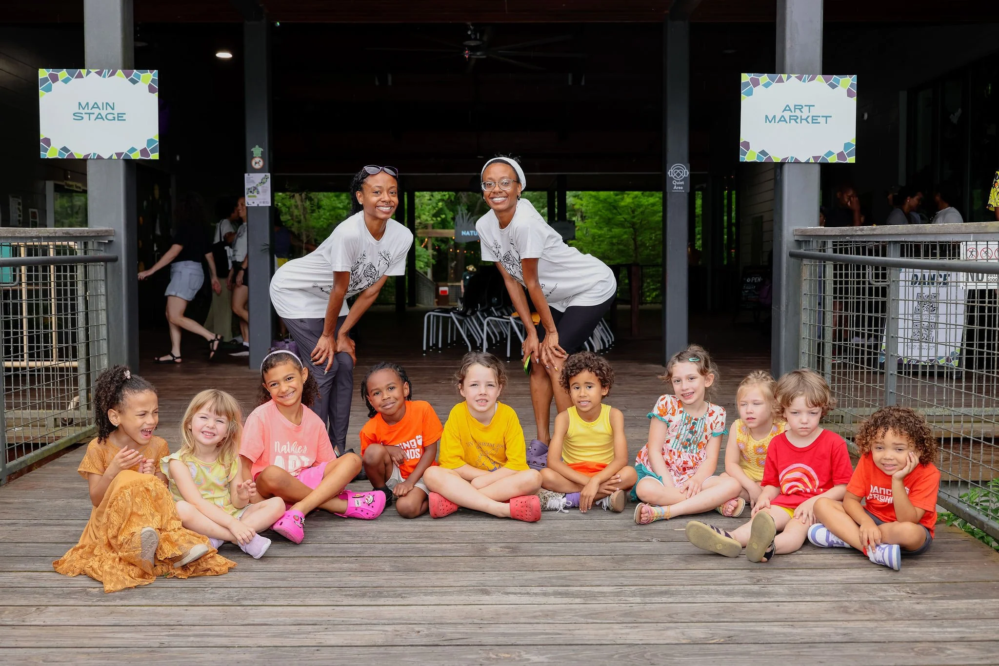 A group of young children sitting on a wooden deck outside, smiling, with two women standing behind them in front of a sign that says 'Main Stage,' at an outdoor event or gathering.