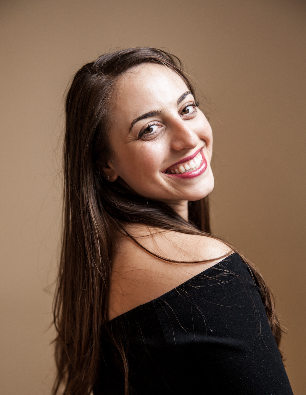 A woman with long brown hair smiling and showing her teeth, wearing a black off-the-shoulder top, against a plain beige background.