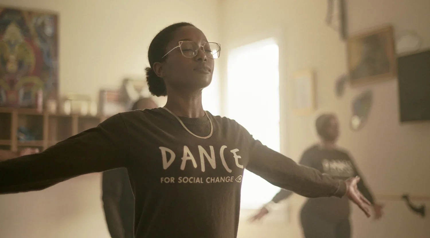 A woman with glasses and a black shirt with the words 'DANCE FOR SOCIAL CHANGE' stands with arms extended in a dance pose, inner a room with sunlight streaming in, and a second person mimicking her dance in the background.
