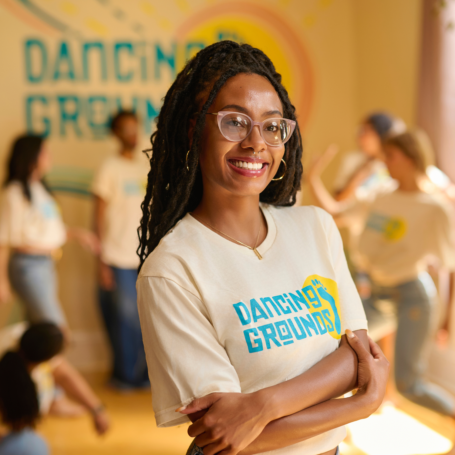 A woman with glasses and dreadlocks smiling with crossed arms, in front of a group of people at a dance event called Dancing Grounds.