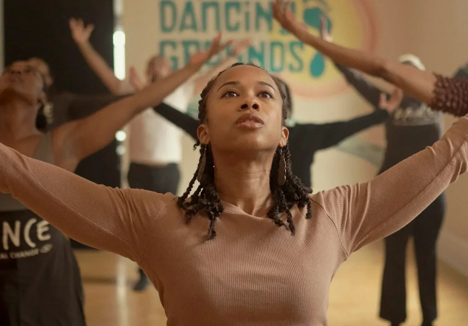 A woman with braids with her arms outstretched in a dance class with other women in the background, all raising their arms, in front of a sign that reads 'Dancing Grannies'.