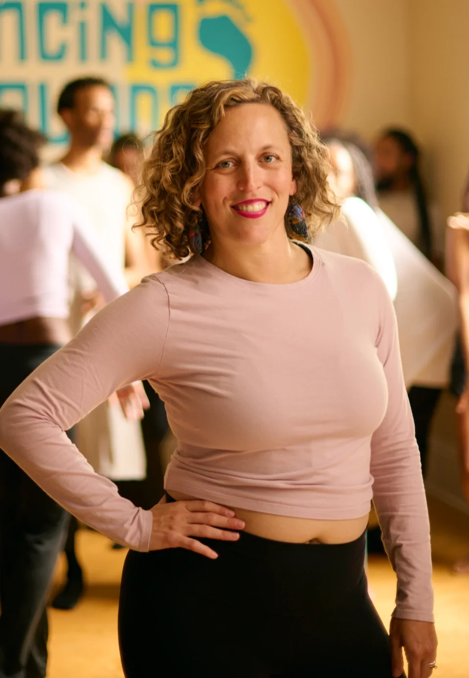 A woman with curly hair smiling, wearing a light pink long-sleeve shirt and black pants, standing in a room with a group of people in the background, and a colorful sign on the wall.
