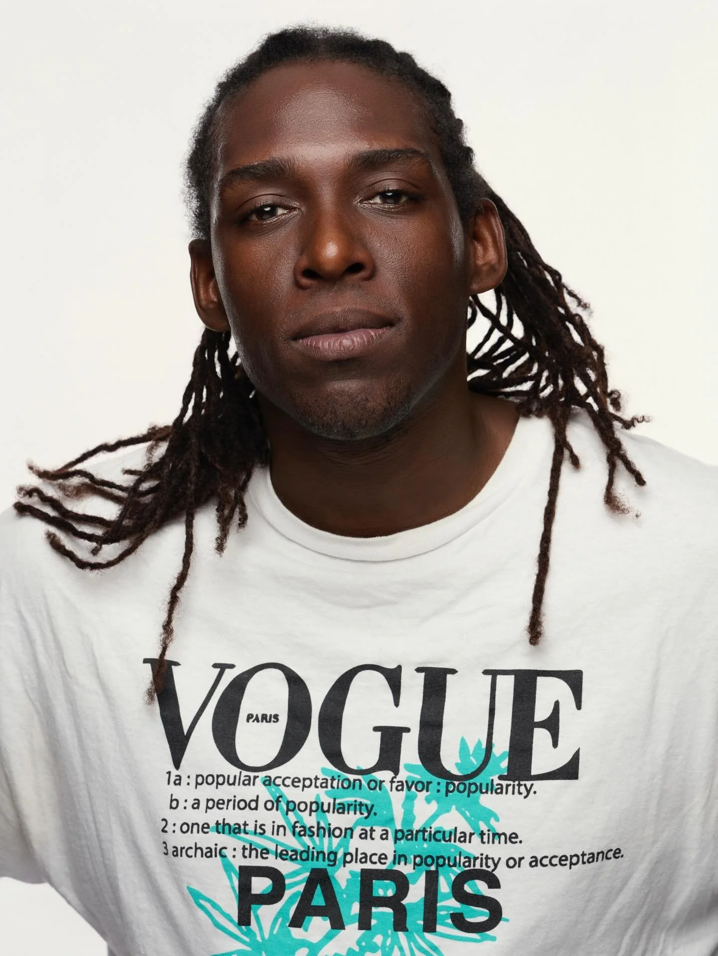 A person with dark skin and dreadlocks wearing a white t-shirt that has black text and a blue graphic design. The photo is a close-up portrait with neutral background.