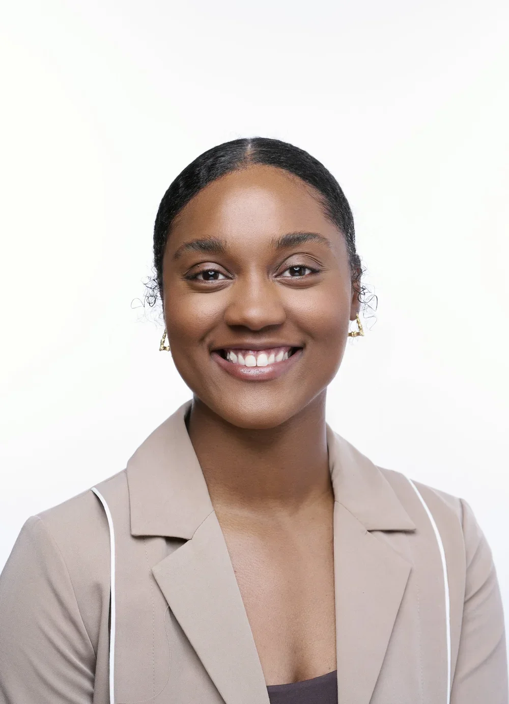A young woman with dark hair pulled back, wearing earrings and a beige blazer, smiling against a white background.