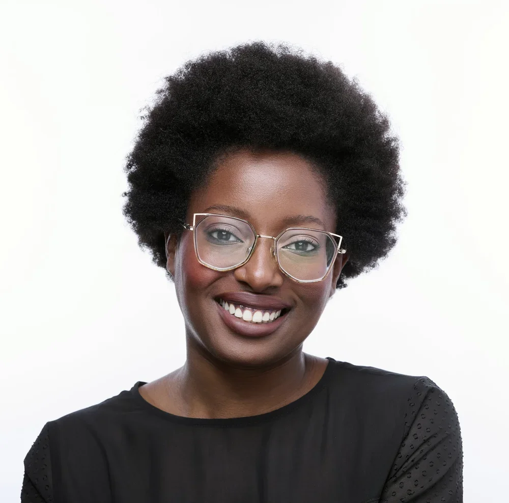 Portrait of a Black woman with natural curly hair, wearing glasses and a black top, smiling against a white background.