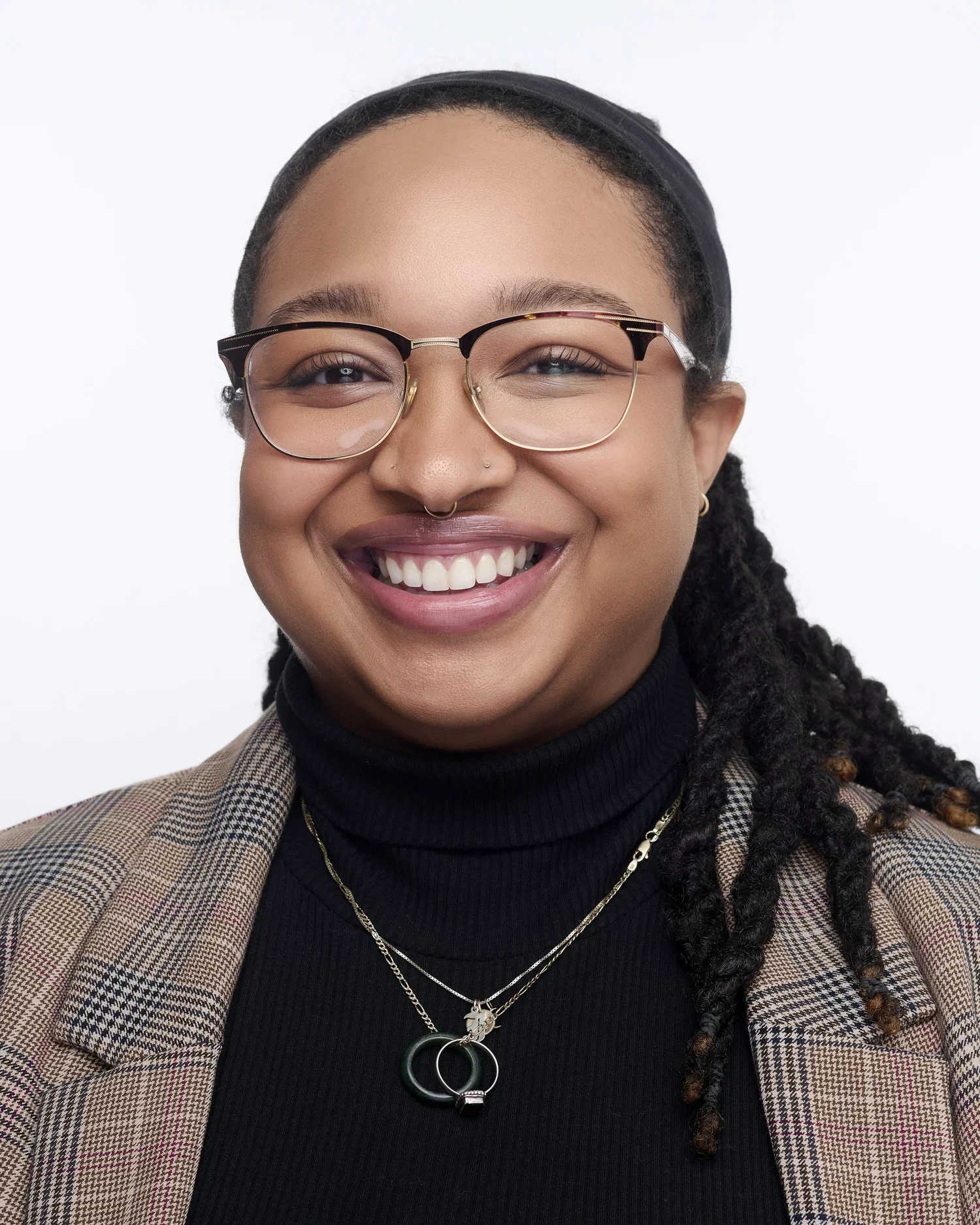 Close-up of a smiling woman with glasses, tattoos, jewelry, and a plaid blazer, isolating her face and upper shoulders against a white background.