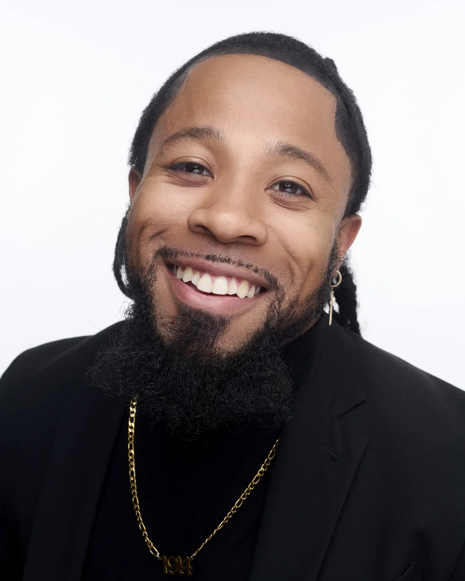 Close-up of a smiling man with a beard and earrings, wearing a black jacket and a gold chain with a pendant that says '194'.