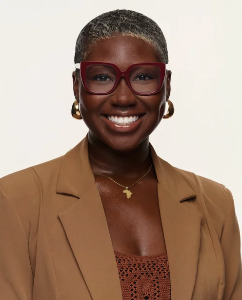 Portrait of a smiling woman with dark skin, short gray hair, glasses, gold earrings, a gold necklace, wearing a tan blazer over a rust-colored top.