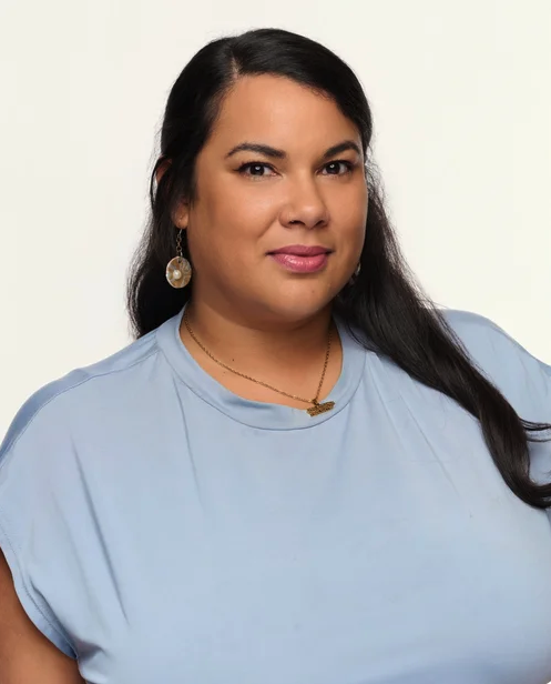 A woman with long dark hair wearing a light blue top, gold earrings, and a gold necklace, looking at the camera with a slight smile.