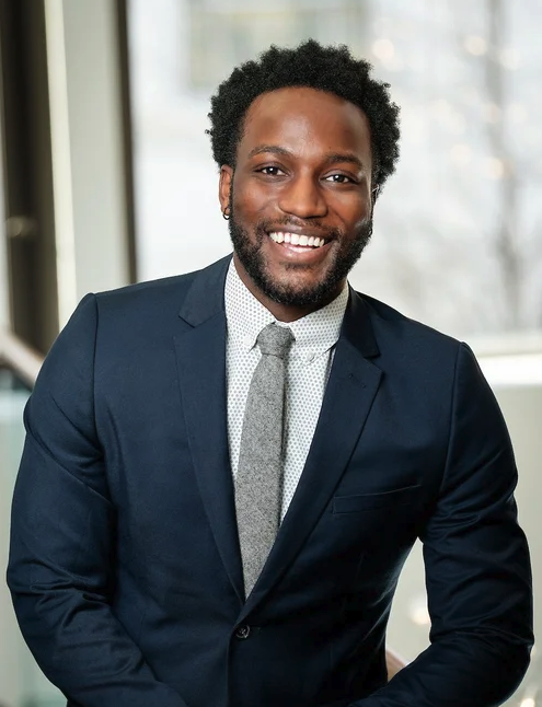 Smiling man in a dark suit and tie, indoor setting with large windows and natural light.