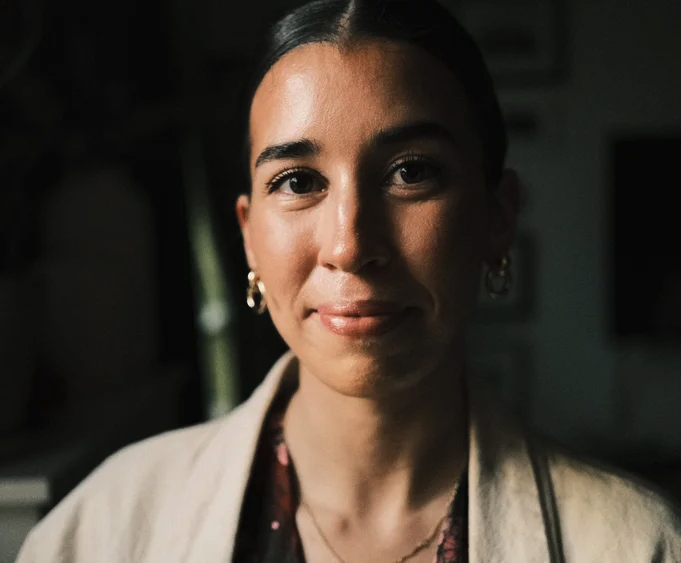 Portrait of a young woman with dark hair, wearing hoop earrings and a beige blazer, smiling softly indoors.