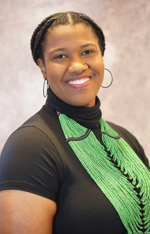 Smiling woman with braided hair, wearing a black top, green beaded necklace, and hoop earrings.