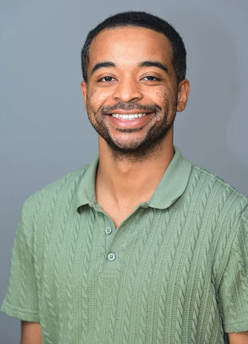 Portrait of a smiling man with short black hair and a light beard, wearing a light green knitted polo shirt, against a gray background.