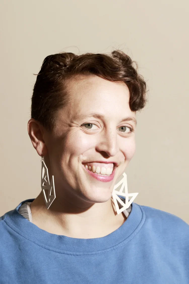 A person with short brown hair smiling, wearing large geometric earrings and a blue shirt against a plain background.