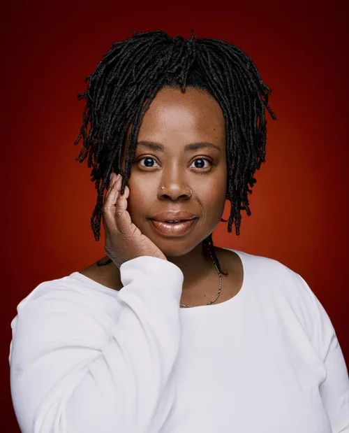 A woman with short dreadlocks, wearing a white long-sleeve shirt, touching her face softly, against a red background.