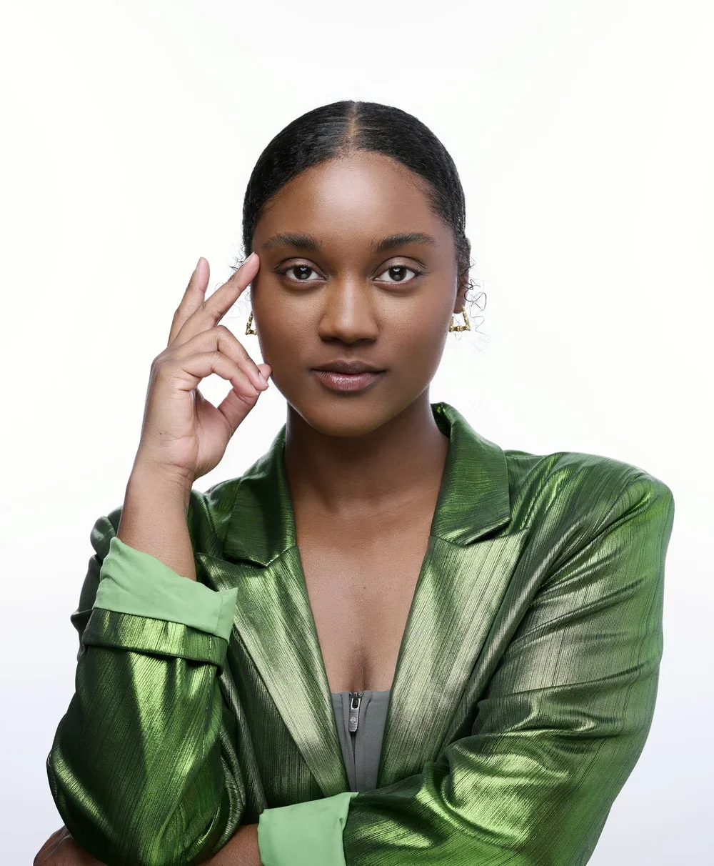 A young African American woman wearing a shiny green jacket with a zipper, touching her temple and looking confidently at the camera against a plain white background.