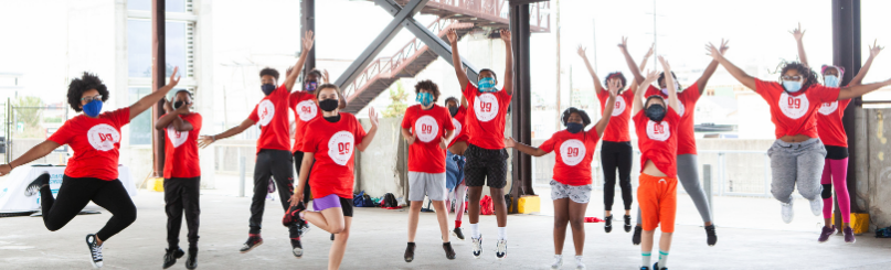 A group of children wearing red shirts and face masks, jumping and raising their hands together in an indoor space with large windows and metal beams.