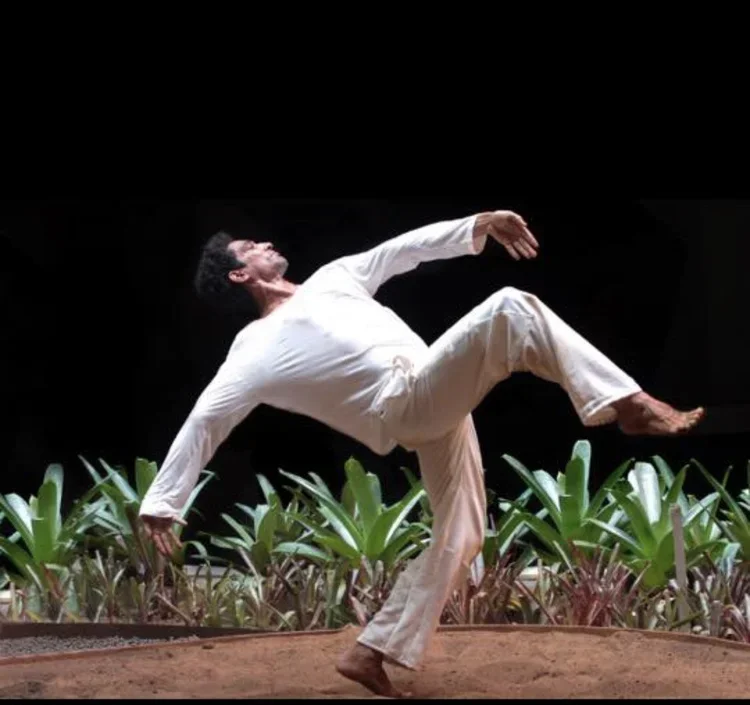 Person in white clothing performing a dance move on a stage with green plants in the background.