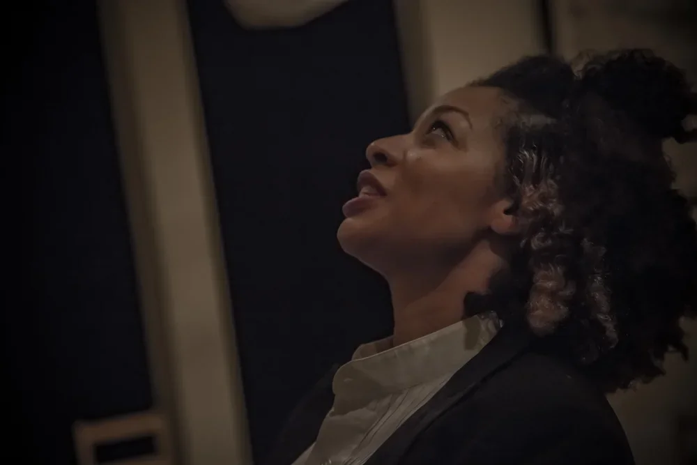 A woman with curly hair looking up, wearing a beige shirt with a dark blazer, in a dimly lit setting.