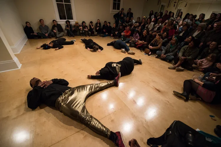 Performers lying on the floor in front of an audience in a dance studio.