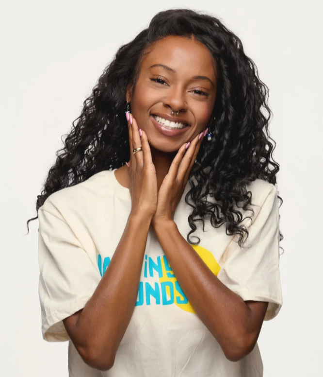 Smiling woman with curly hair, touching her face, wearing a beige T-shirt with blue and yellow text, against a plain white background.