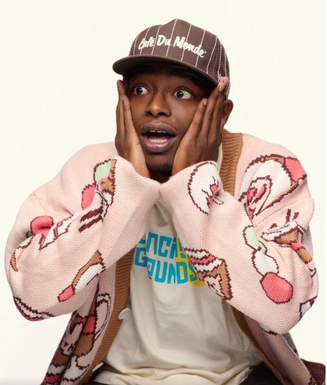A young man with surprised expression, hands on his cheeks, wearing a brown cap with white writing, a pink patterned sweater, and a white T-shirt.