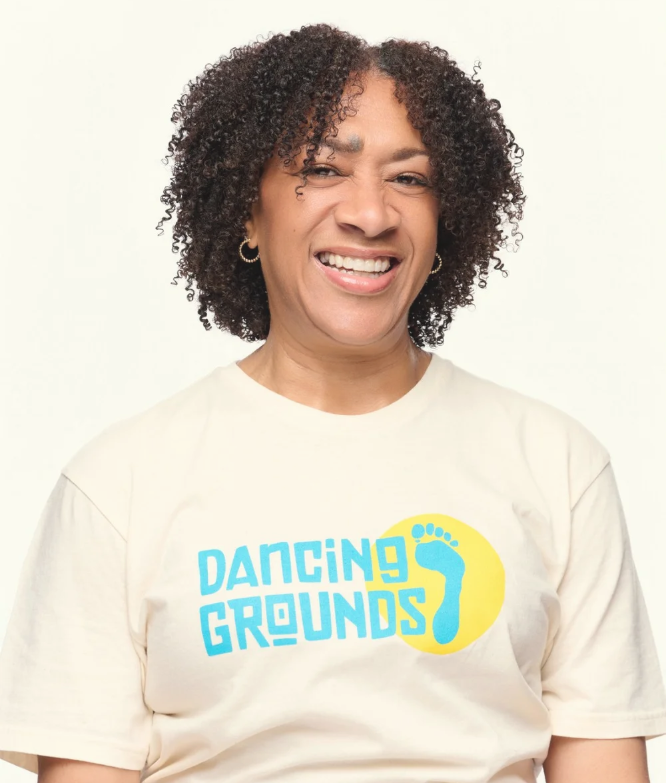 A woman smiling, wearing a white T-shirt with a logo that reads 'Dancing Grounds' and features a yellow circle with a blue footprint.
