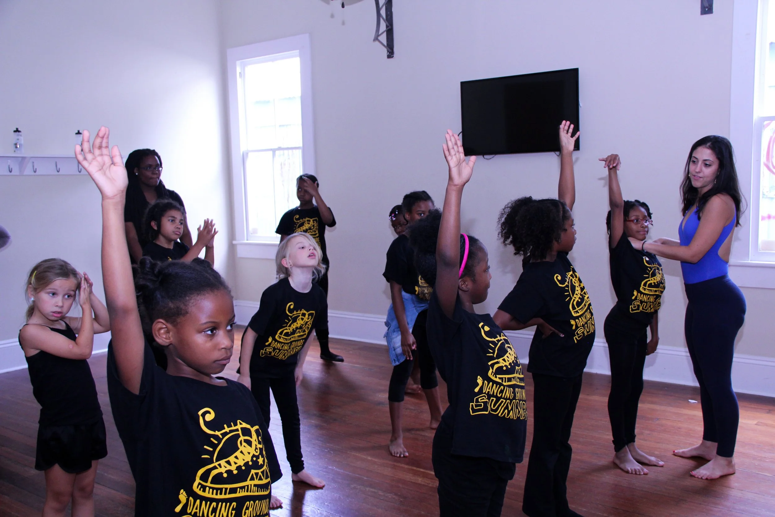 Children practicing dance moves in a dance class, with some raising their hands, in a room with white walls, wooden floors, and large windows.
