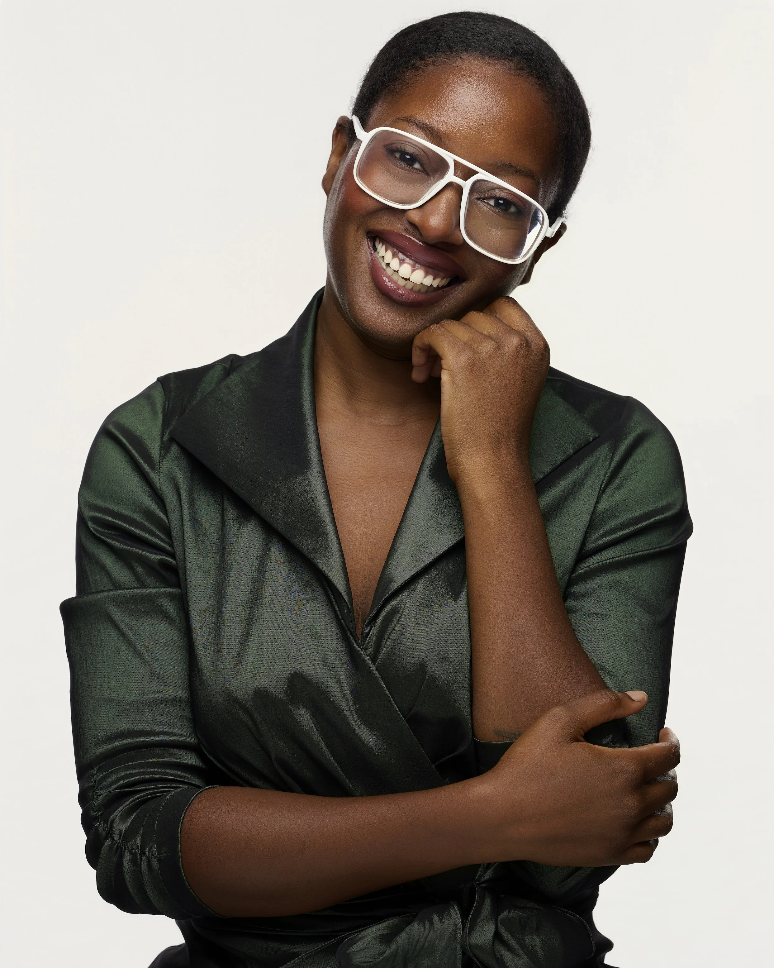 Smiling woman with short hair wearing transparent glasses and a dark satin blazer, sitting against a white background.