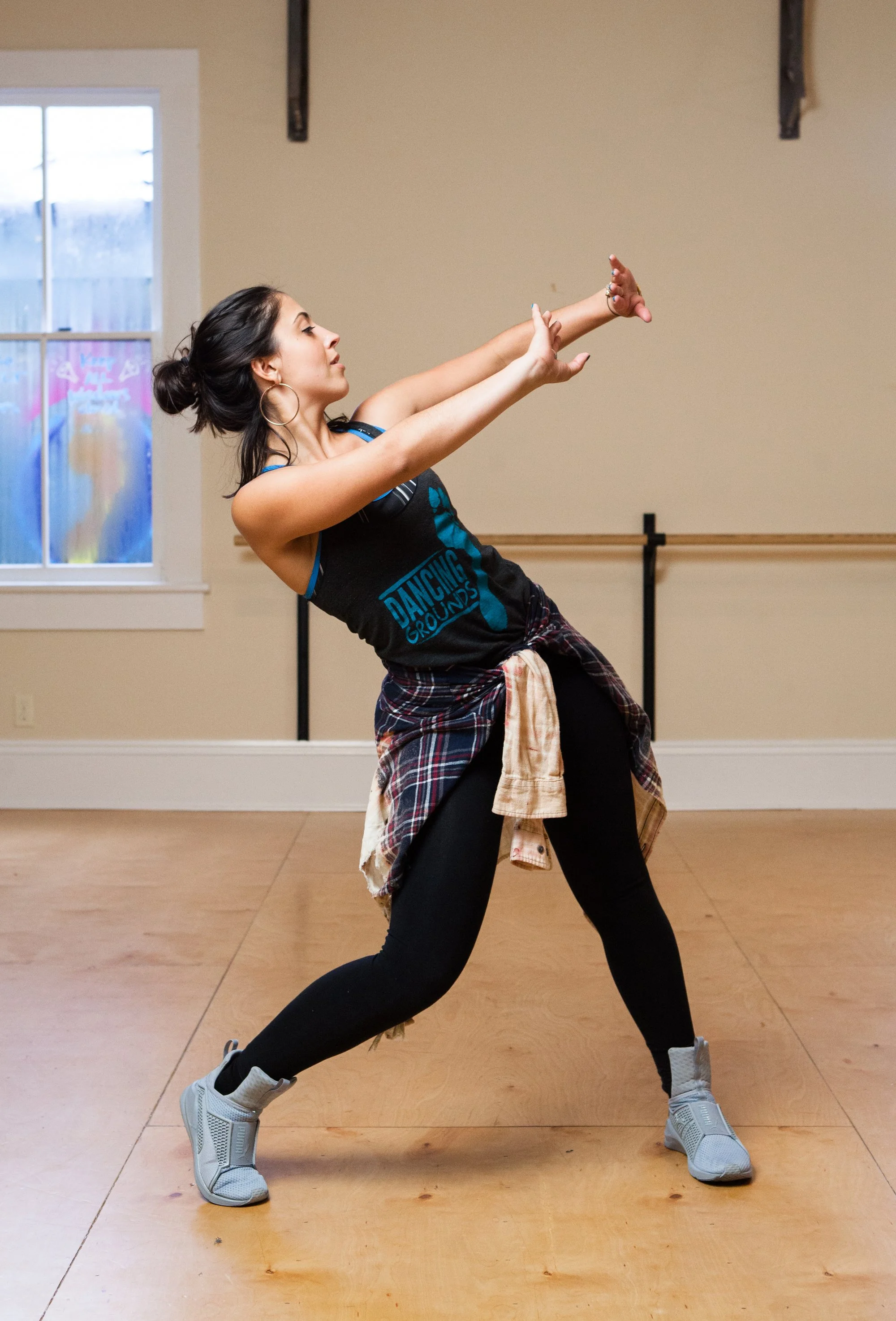 A woman dancing in a dance studio, wearing a t-shirt that says 'DANCING GROUNDS', black leggings, gray sneakers, and a plaid shirt tied around her waist, with her arms extended and in a dynamic pose.