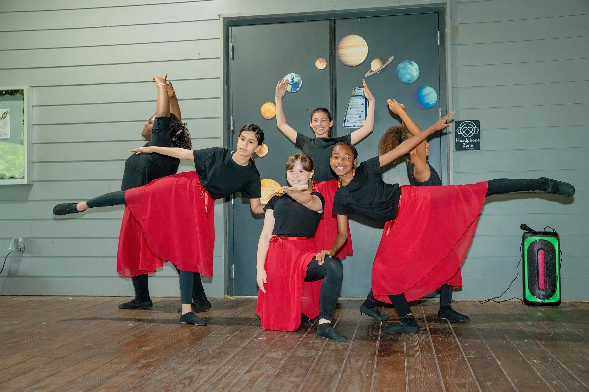 Five young girls performing a dance routine on a wooden stage, wearing black shirts and red skirts, with a space-themed wall decoration behind them. One girl in the center is smiling with arms raised, while others pose in various ballet positions.