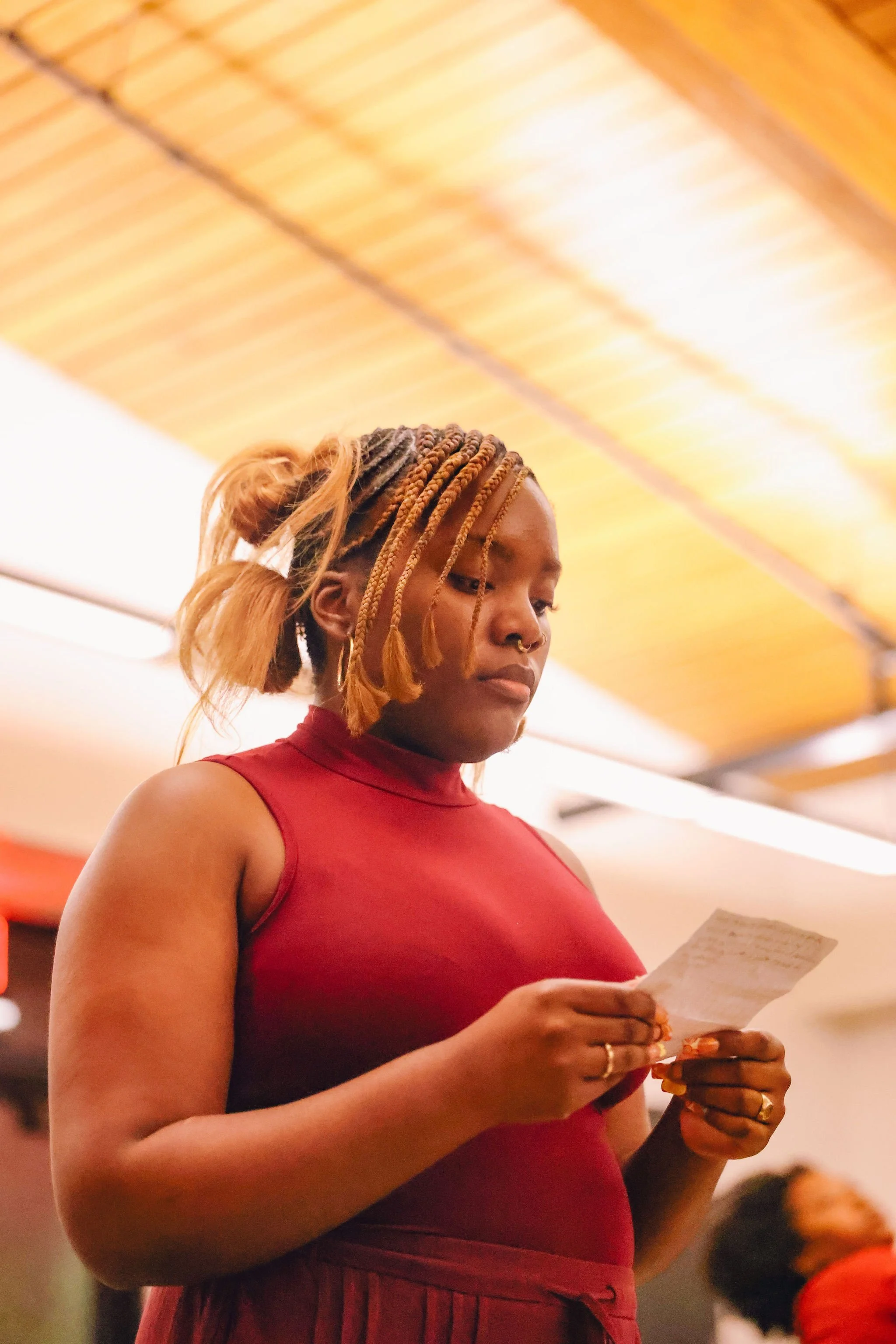 A woman with braided hair and a nose ring, wearing a sleeveless red top, is reading a piece of paper in a warmly lit indoor setting.