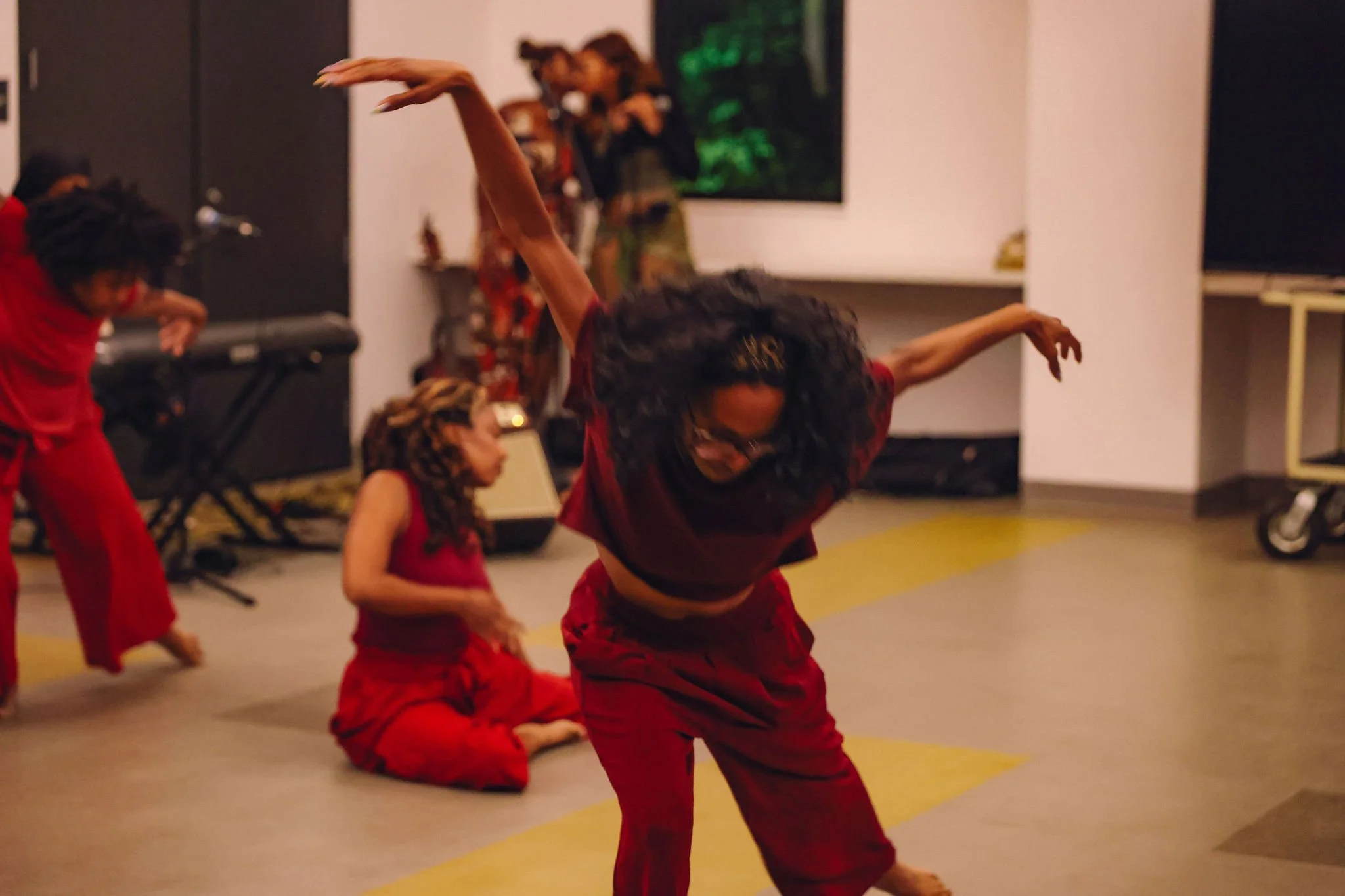 People practicing dance or martial arts in a room, wearing red costumes, with some standing and one person kneeling on the floor.