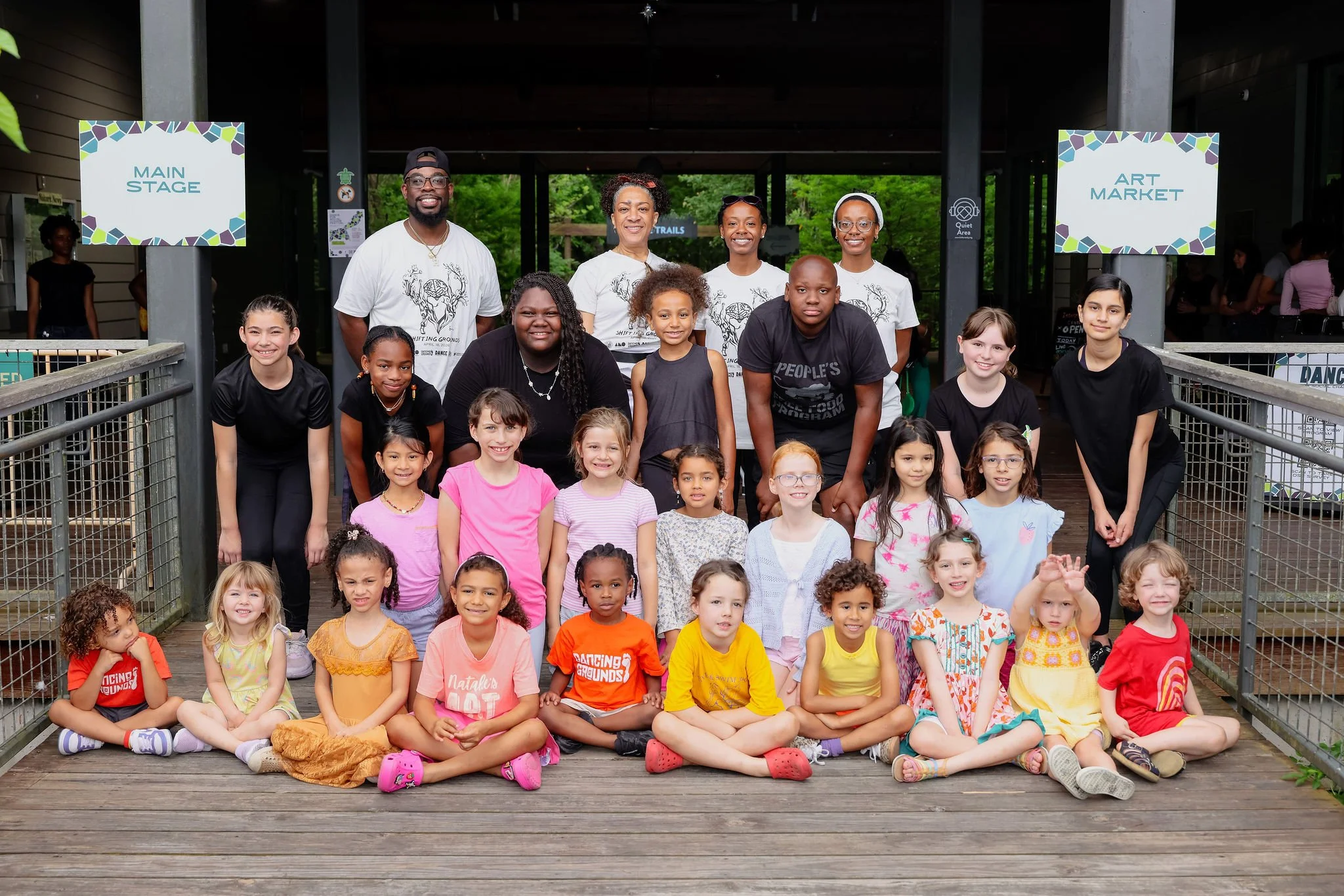 A large group of children and adults posing on a wooden deck in front of a building with signs indicating main stage and art market. The children are sitting and standing, smiling and facing the camera, with the adults standing behind them, also smiling. The scene is outdoors with green trees in the background.