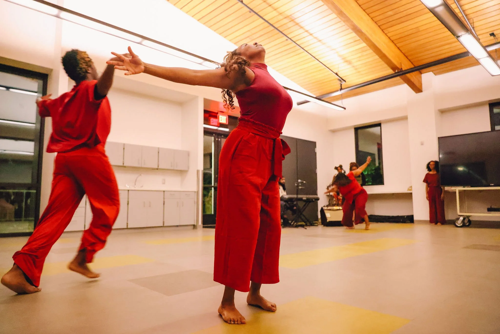 A woman in a red top and red pants practicing yoga in a studio with children, with her arms stretched out and head tilted back, while children in red outfits perform various yoga poses in the background.