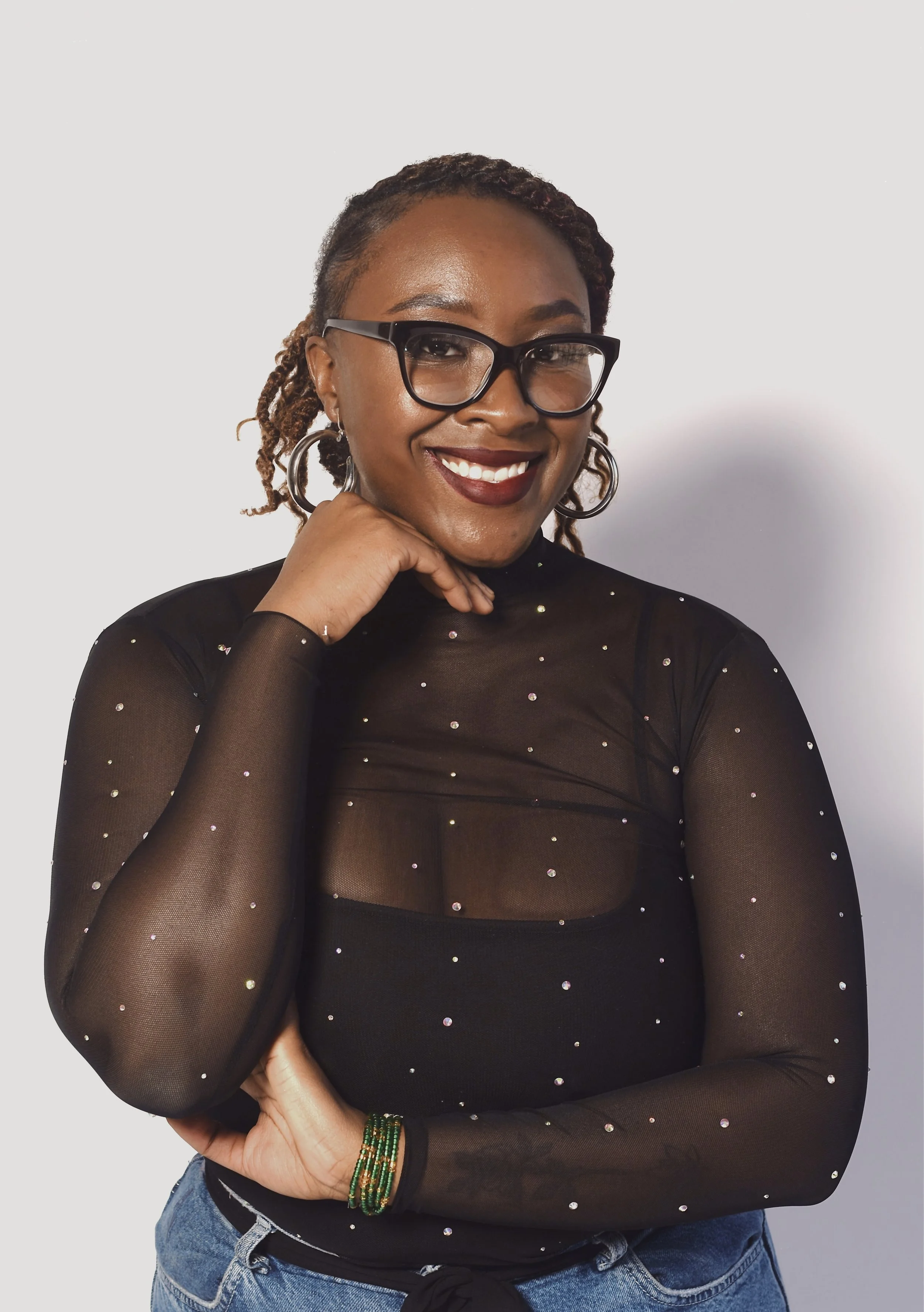 A woman smiling, wearing black glasses, hoop earrings, a black mesh top with glittering dots, and blue jeans, standing against a light background.