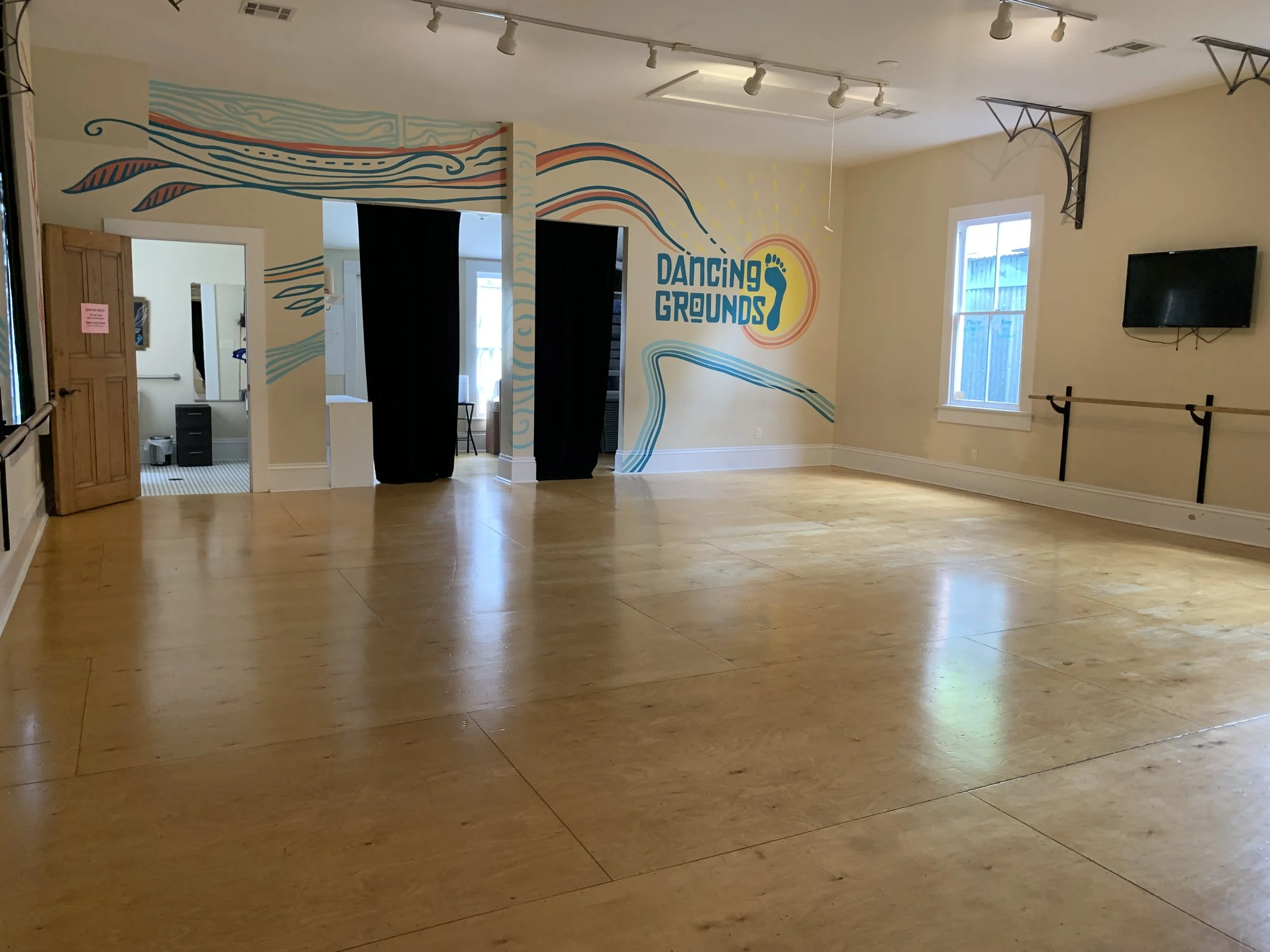 An interior dance studio with wooden floors, a colorful mural on the wall, and a sign that reads 'Dancing Grounds' with a foot graphic. There are windows, a TV, and dance barres.