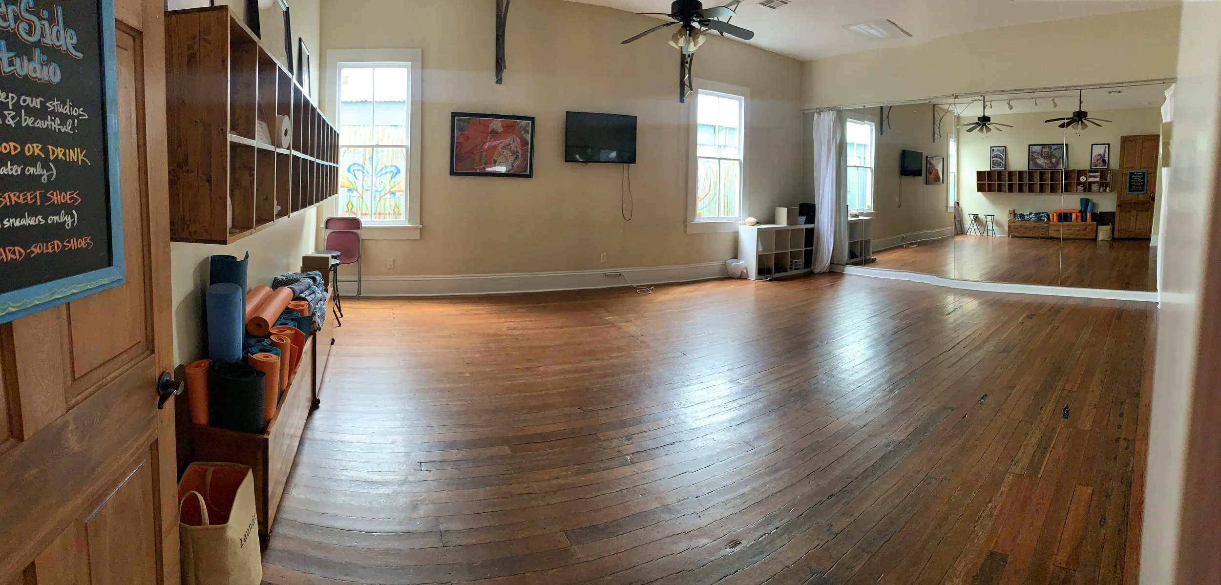 An empty room with hardwood floors, beige walls, several windows, wall-mounted TV, mirrored wall, ceiling fans, and storage shelves, likely a yoga or dance studio.
