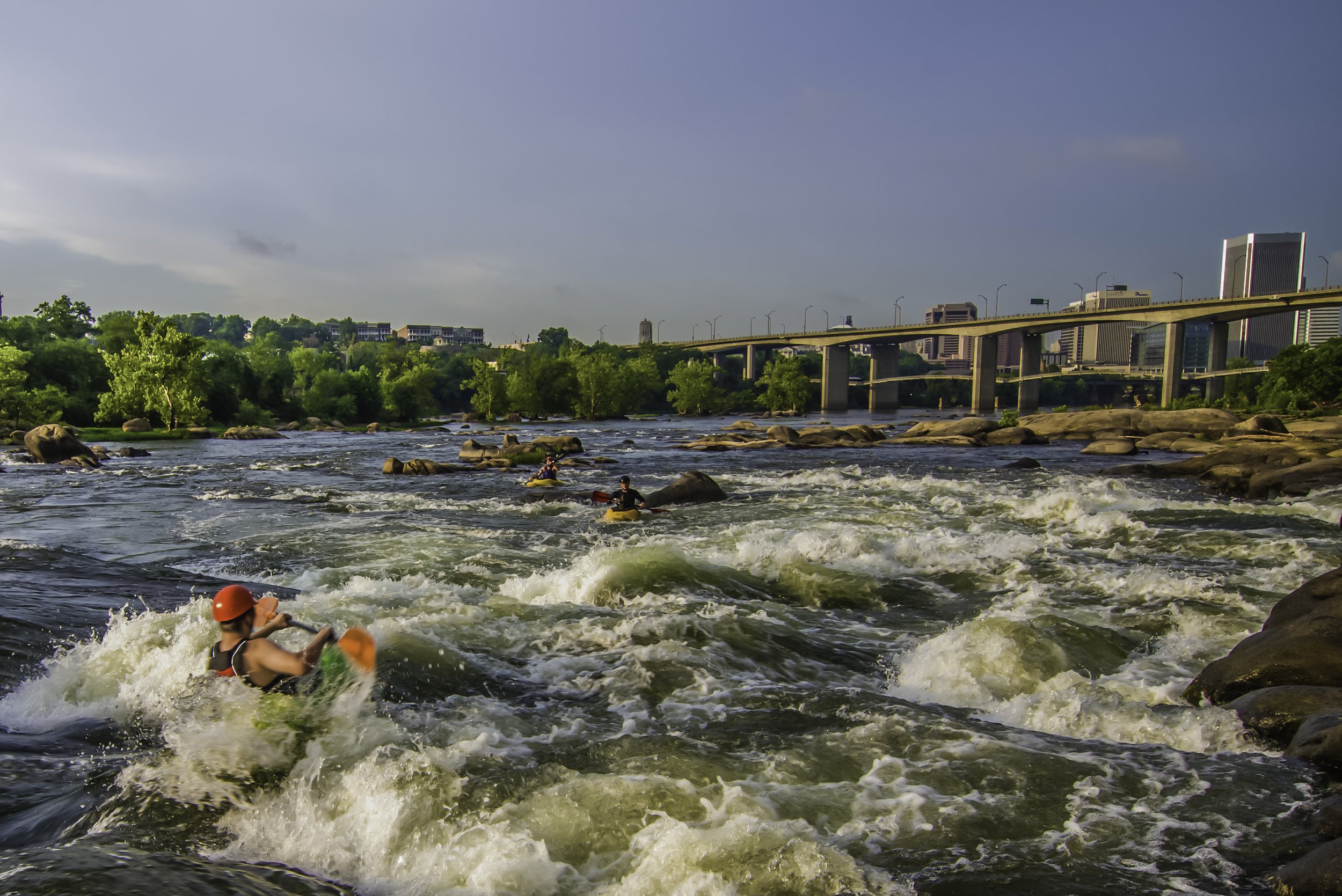 People kayaking on a river with city buildings and a bridge in the background.