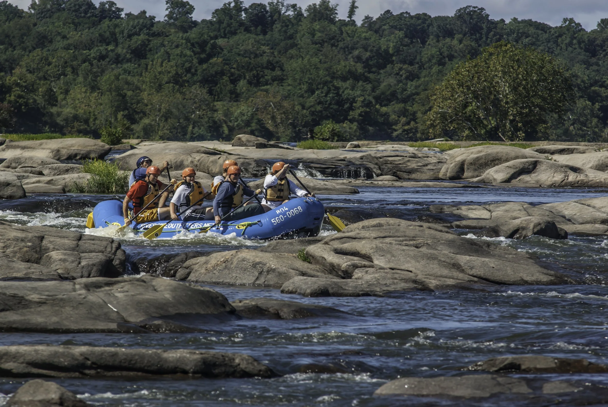 A group of six people white water rafting on a river surrounded by rocks and lush green trees with a blue sky overhead.