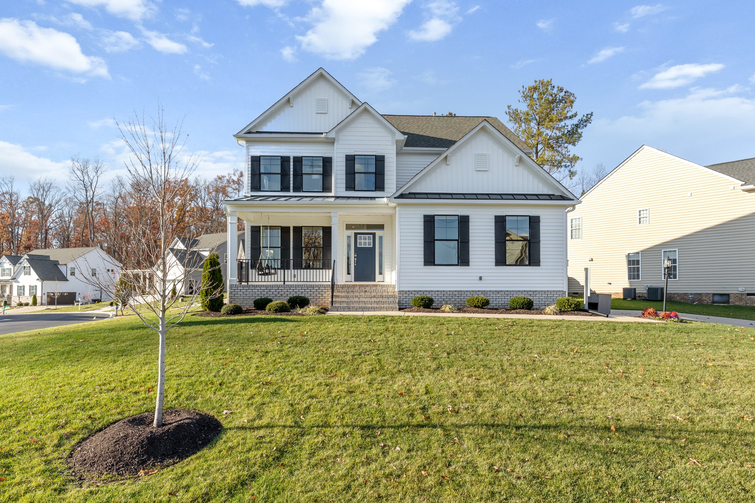 Front view of a white two-story house with black window shutters, a porch, and a front yard with green grass and a small tree.