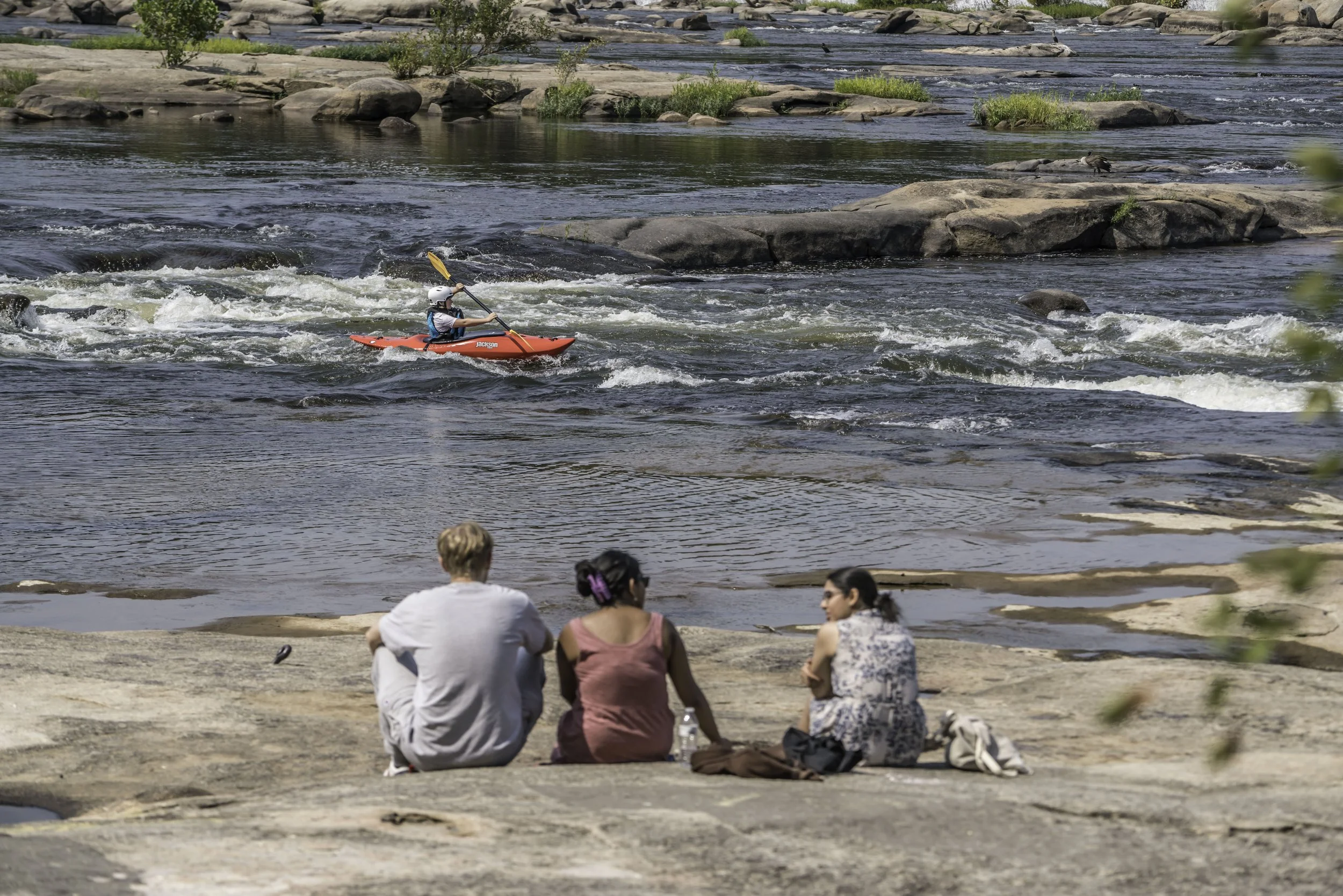 A person kayaking in a river with rocky surroundings, while several people sit on the riverbank watching.