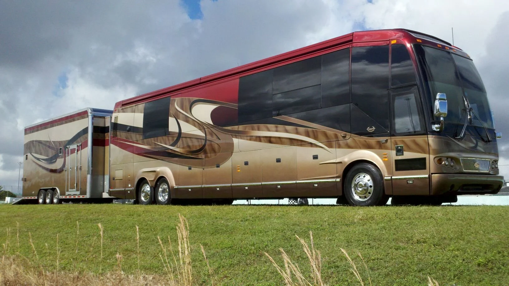 Large gold and black luxury motorhome with artistic swirl patterns parked on a grassy field, behind a trailer, under partly cloudy skies.