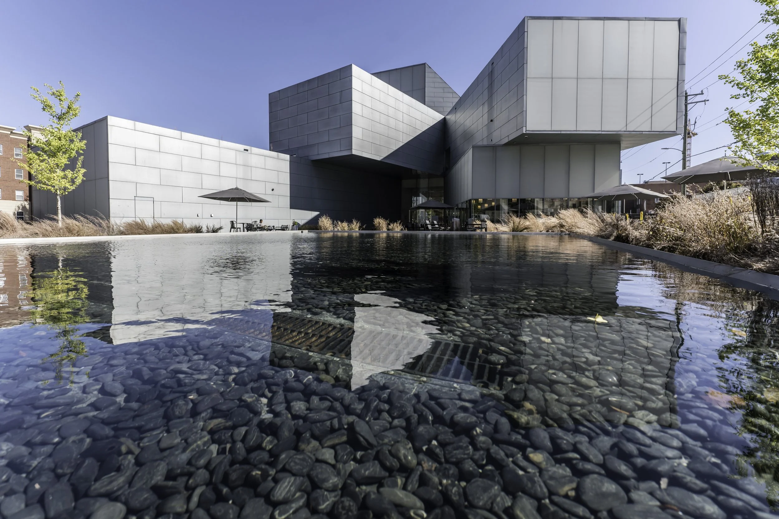 Modern building with gray exterior, outdoor seating, trees, and a reflecting pool in the foreground under a clear blue sky.