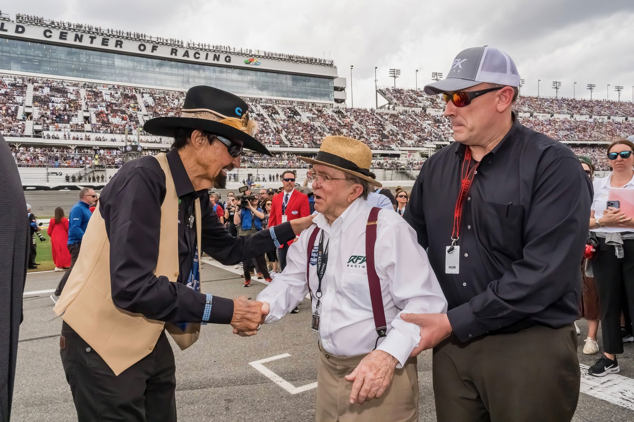Three men shaking hands at a racetrack with a crowded grandstand in the background.