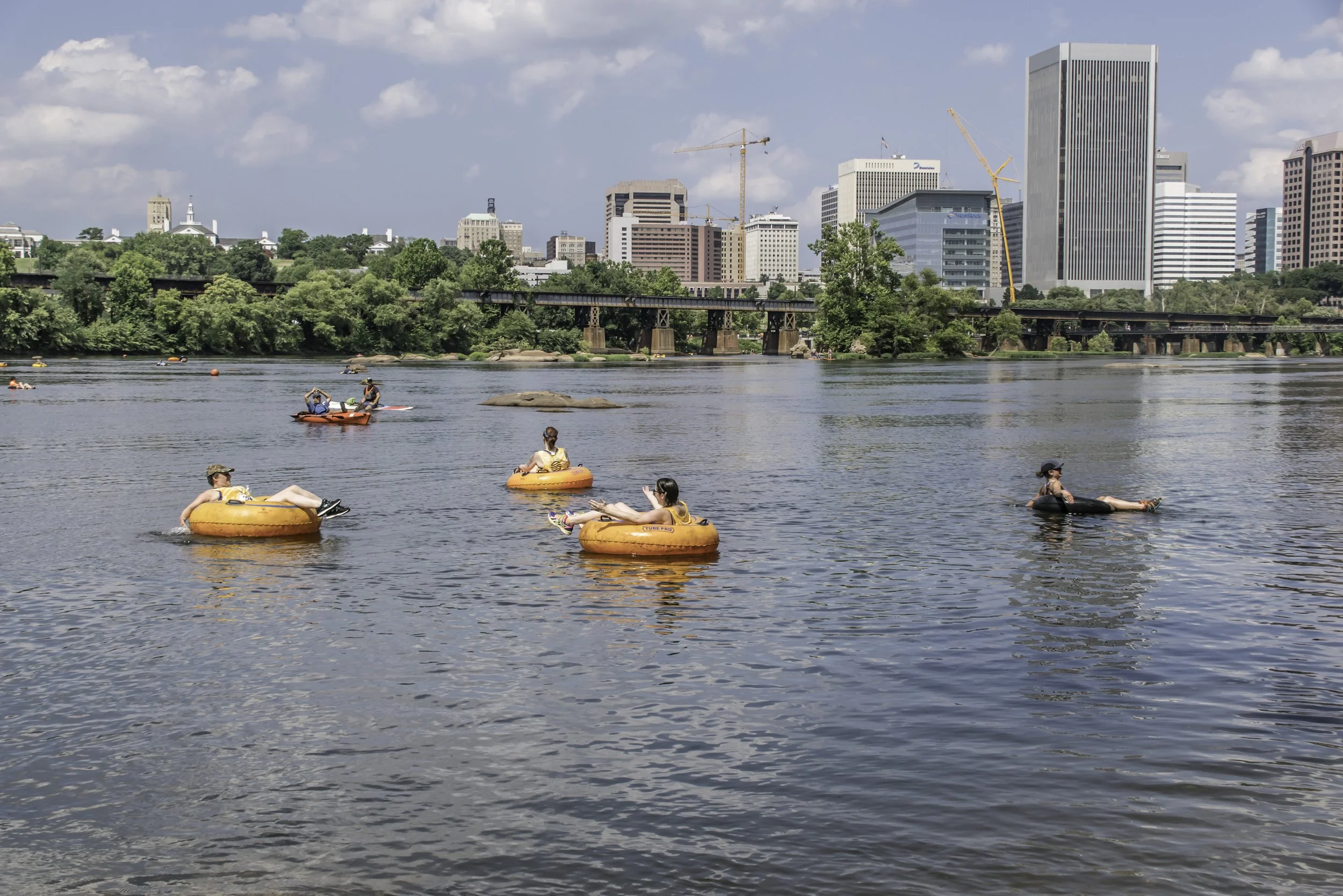People floating on the water on orange inner tubes in front of a city skyline with tall buildings and construction cranes.