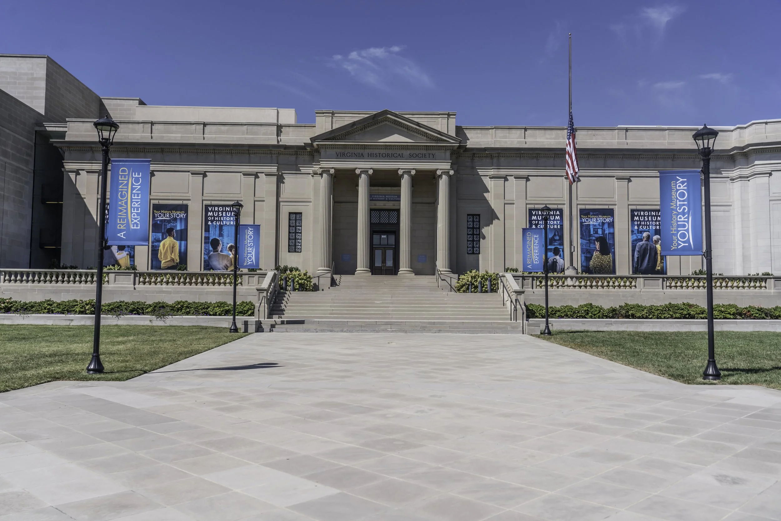 The Virginia Historical Society building with steps leading up to the entrance, flanked by three lampposts with banners that read "A REIMAGINED EXPERIENCE" and "YOUR STORY." The building has classical columns and an American flag flying in front. The sky is clear and blue.