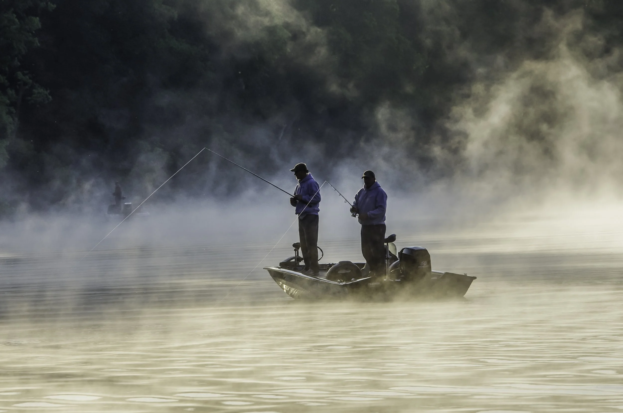 Two men fishing from a small boat on a misty lake at dawn, with lush green trees in the background.